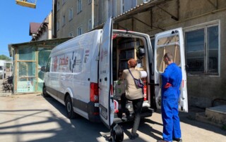 A transporter in front of a industry building with two people standing at the opened back door of the car.