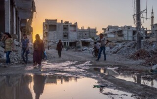 Several people walk across muddy streets in the middle of destroyed buildings.